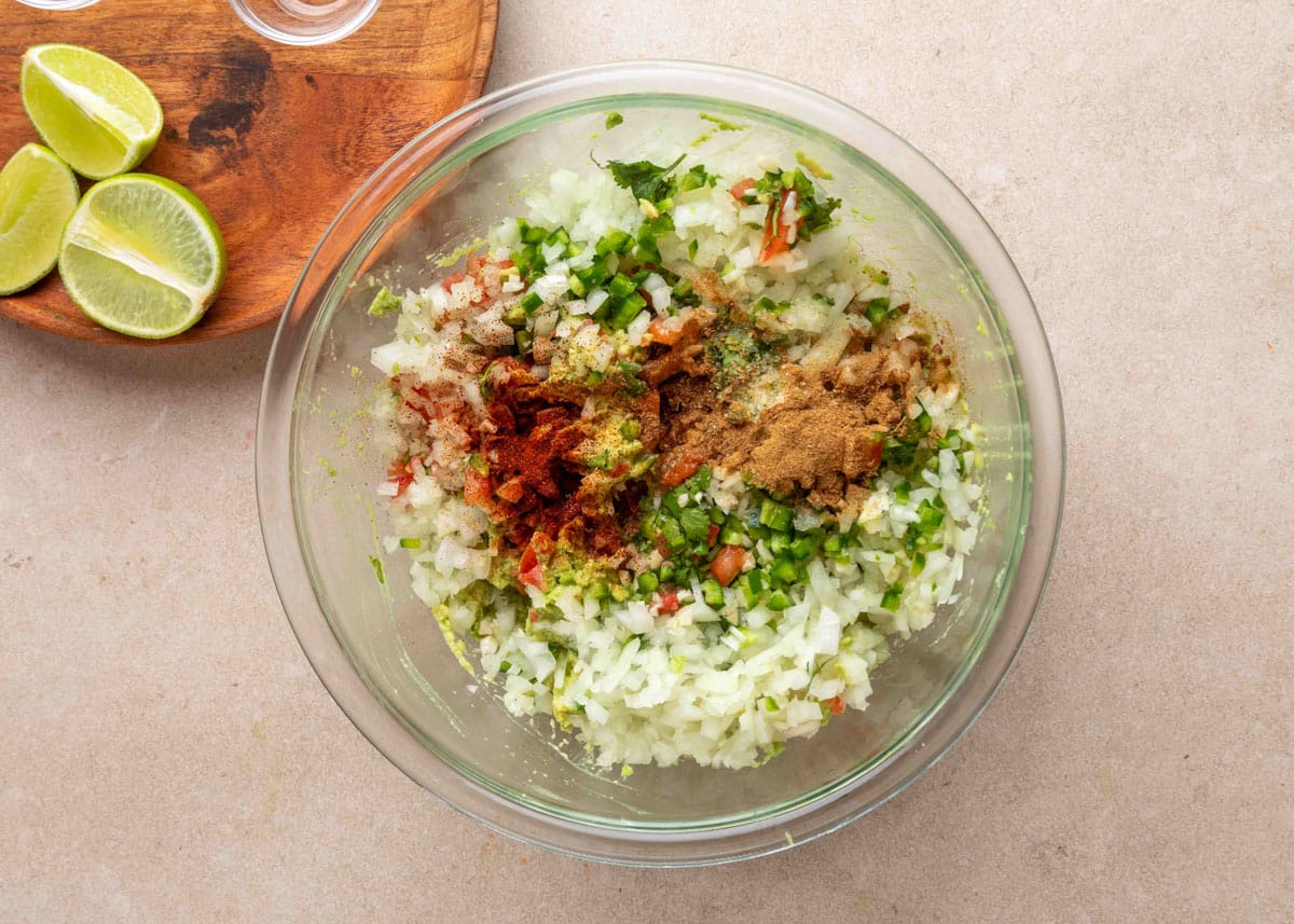 Chopped onions, jalape&ntilde;os, tomatoes, and cilantro in a glass bowl topped with ground cumin, chipotle powder, and other spices, ready to be mixed into guacamole; lime wedges are visible on a wooden board nearby.
