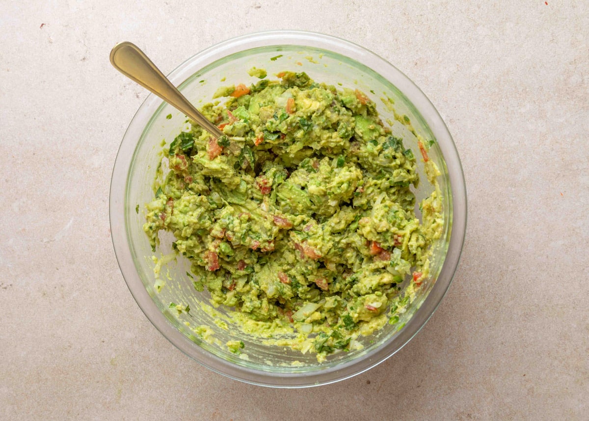 Fully mixed chunky guacamole in a glass bowl with a fork, showing visible pieces of avocado, tomato, onion, jalape&ntilde;o, and cilantro against a neutral background.