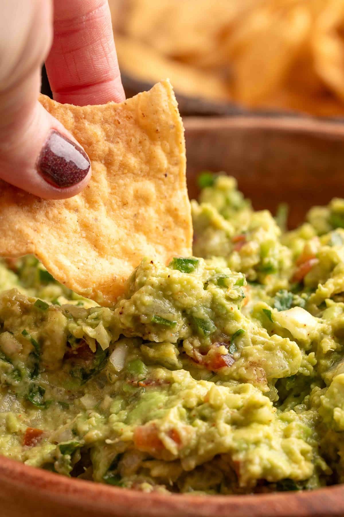 A hand holding a tortilla chip dipping into a rustic bowl of chunky guacamole with visible onion, tomato, and cilantro.
