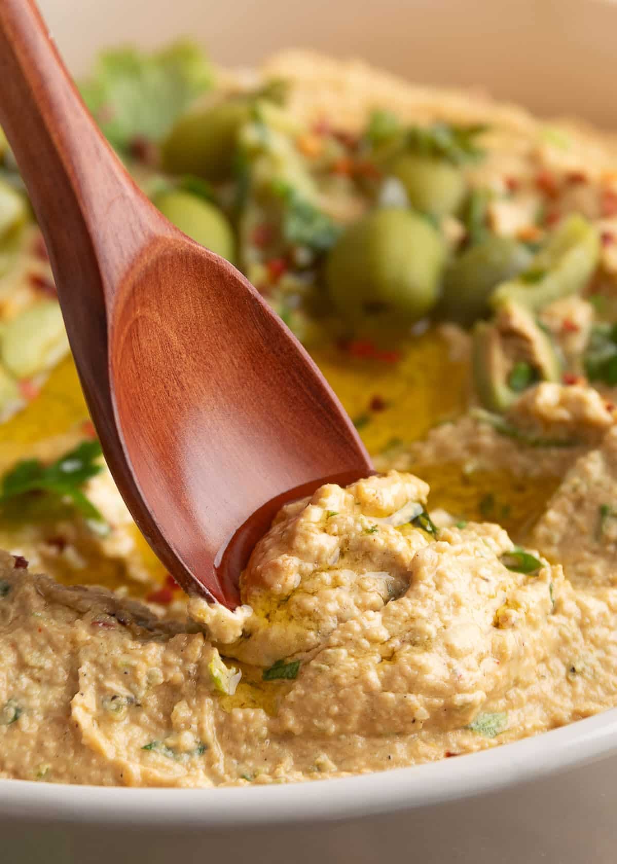 Close-up of a wooden spoon lifting a scoop of creamy green olive hummus from a bowl, with chopped herbs and olives visible in the background.