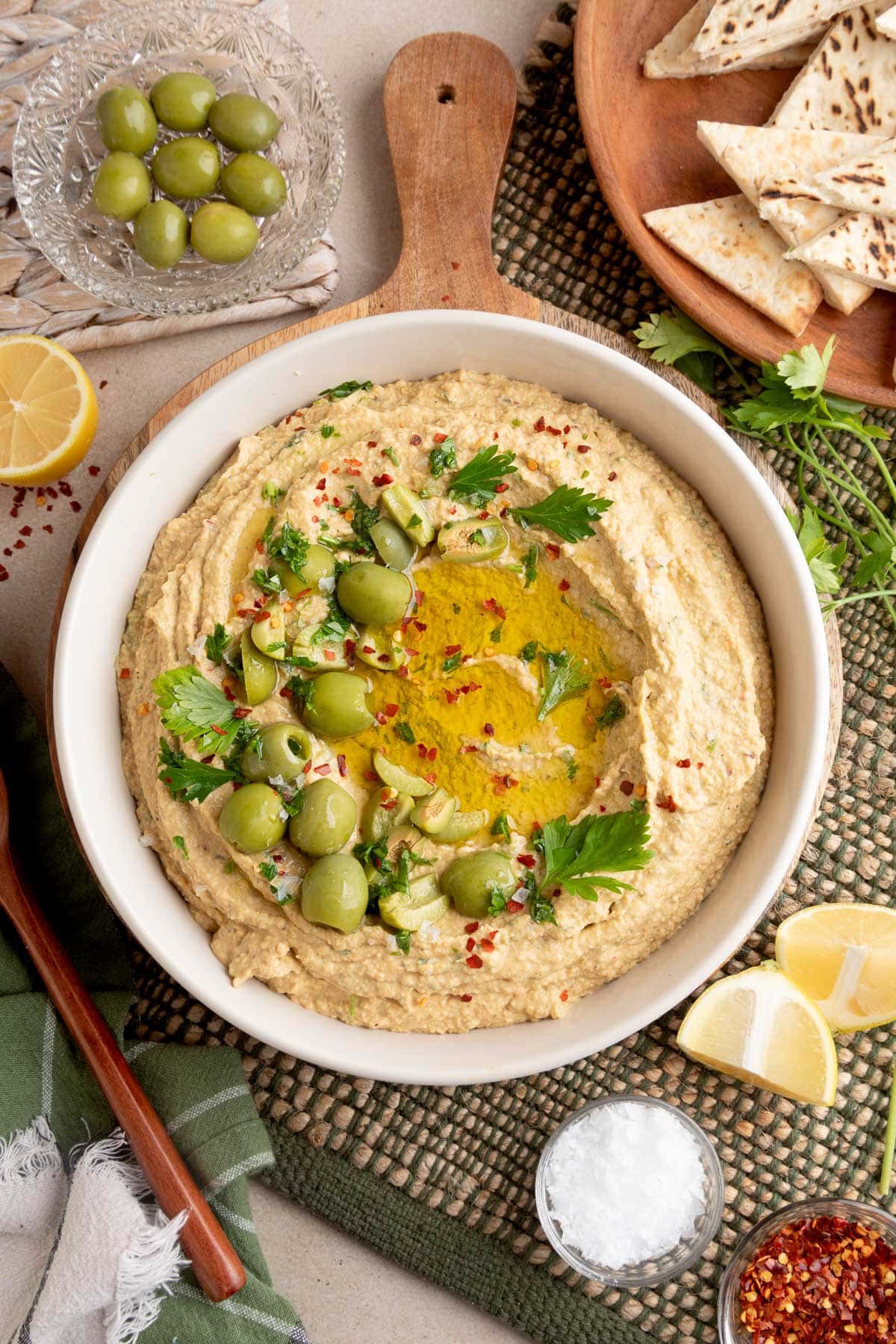 Overhead view of a bowl of green olive hummus topped with chopped olives, parsley, olive oil, and red pepper flakes, surrounded by pita wedges, lemon wedges, and small dishes of garnishes on a textured surface.