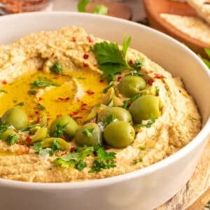 Close-up of creamy green olive hummus in a white bowl, topped with whole and sliced green olives, chopped parsley, red pepper flakes, and a drizzle of olive oil.