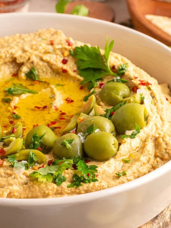 Close-up of creamy green olive hummus in a white bowl, topped with whole and sliced green olives, chopped parsley, red pepper flakes, and a drizzle of olive oil.