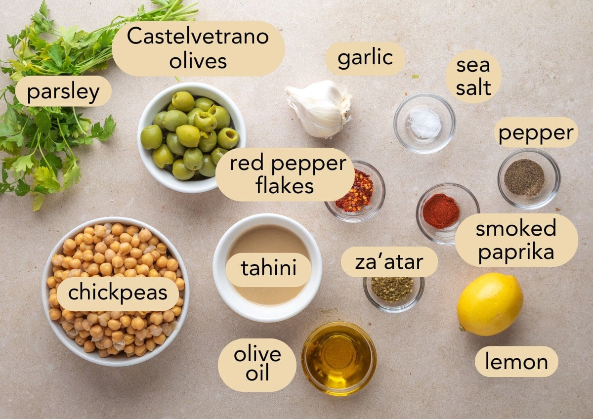 Labeled flat lay of ingredients for green olive hummus on a beige surface, including Castelvetrano olives, chickpeas, tahini, olive oil, lemon, parsley, garlic, sea salt, pepper, smoked paprika, za&rsquo;atar, and red pepper flakes in small bowls.