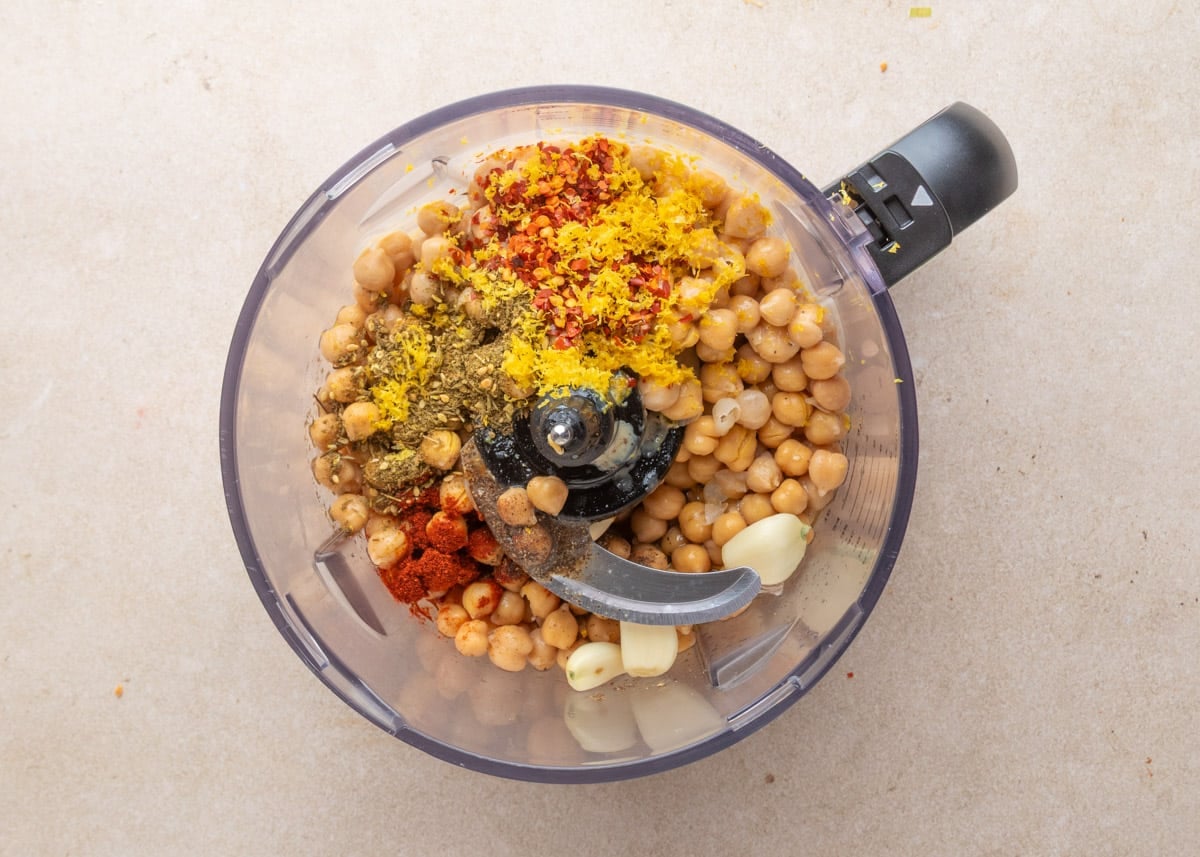 Unblended chickpeas in a food processor bowl with whole garlic cloves, lemon zest, red pepper flakes, za&rsquo;atar, smoked paprika, black pepper, and sea salt arranged on top.
