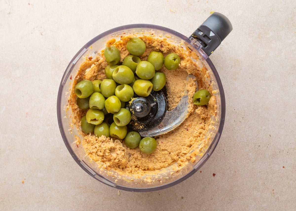 Partially blended hummus in a food processor bowl, topped with whole Castelvetrano olives just before the final blend.