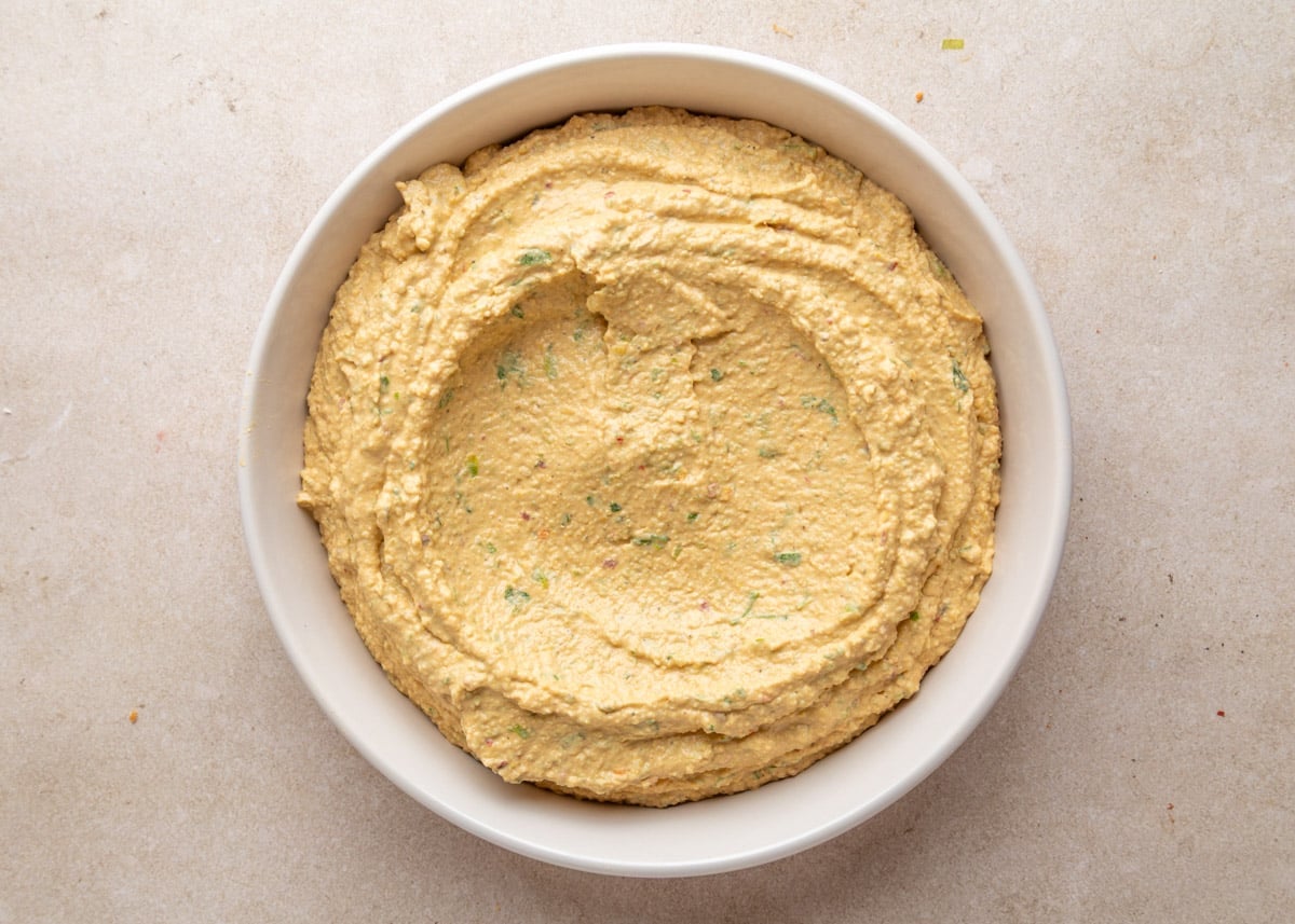 Overhead view of a white bowl filled with creamy green olive hummus, swirled on top and flecked with bits of herbs and finely chopped olives.