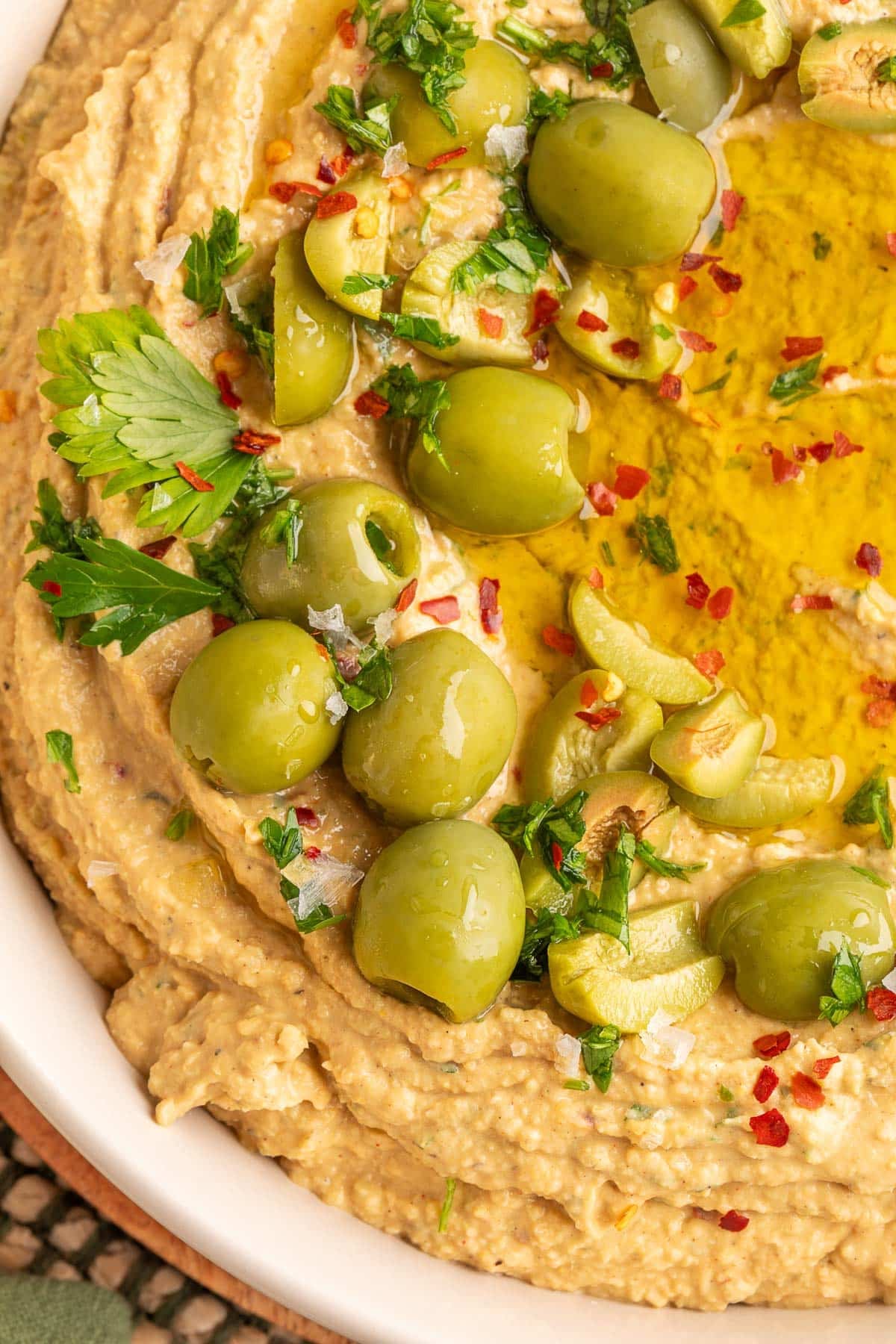 Close-up of creamy green olive hummus swirled in a bowl, topped with whole and sliced green olives, chopped parsley, crushed red pepper flakes, flaky sea salt, and a drizzle of olive oil.