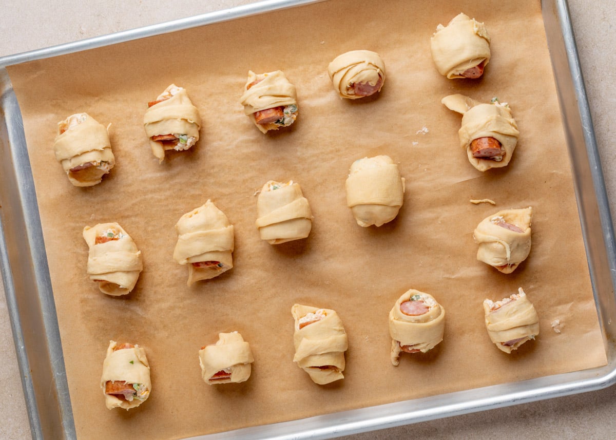 Overhead view of 16 unbaked jalape&ntilde;o popper pigs in a blanket arranged on a parchment-lined sheet pan, each wrapped in crescent dough and filled with smoked sausage and jalape&ntilde;o cream cheese mixture.