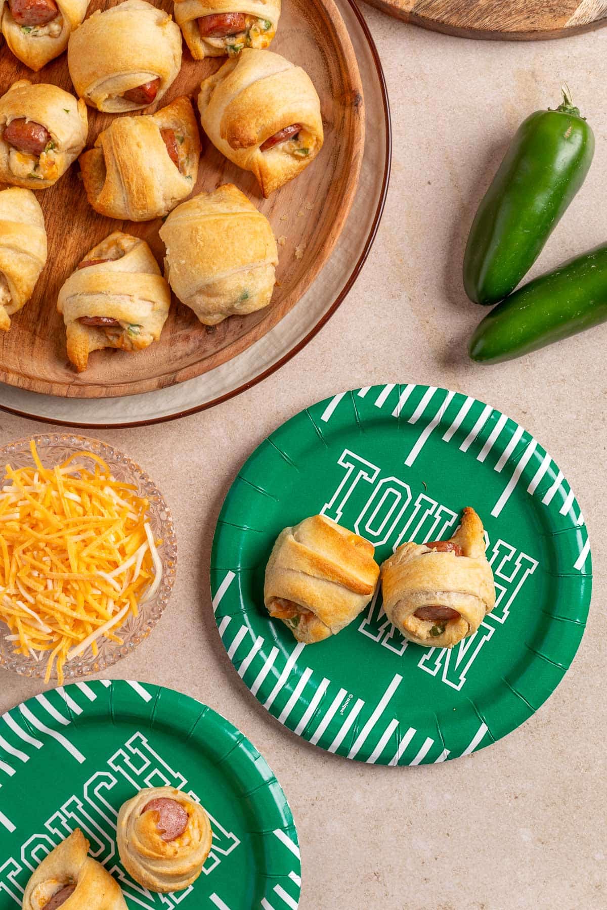 Overhead view of jalape&ntilde;o popper pigs in a blanket served on a wooden platter and green football-themed paper plates, with shredded cheese and fresh jalape&ntilde;os nearby on a beige surface.