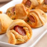 Golden brown jalapeño popper pigs in a blanket arranged on a white rectangular platter, showing melted cheese, sliced sausage, and bits of green jalapeño peeking through the crescent roll dough.