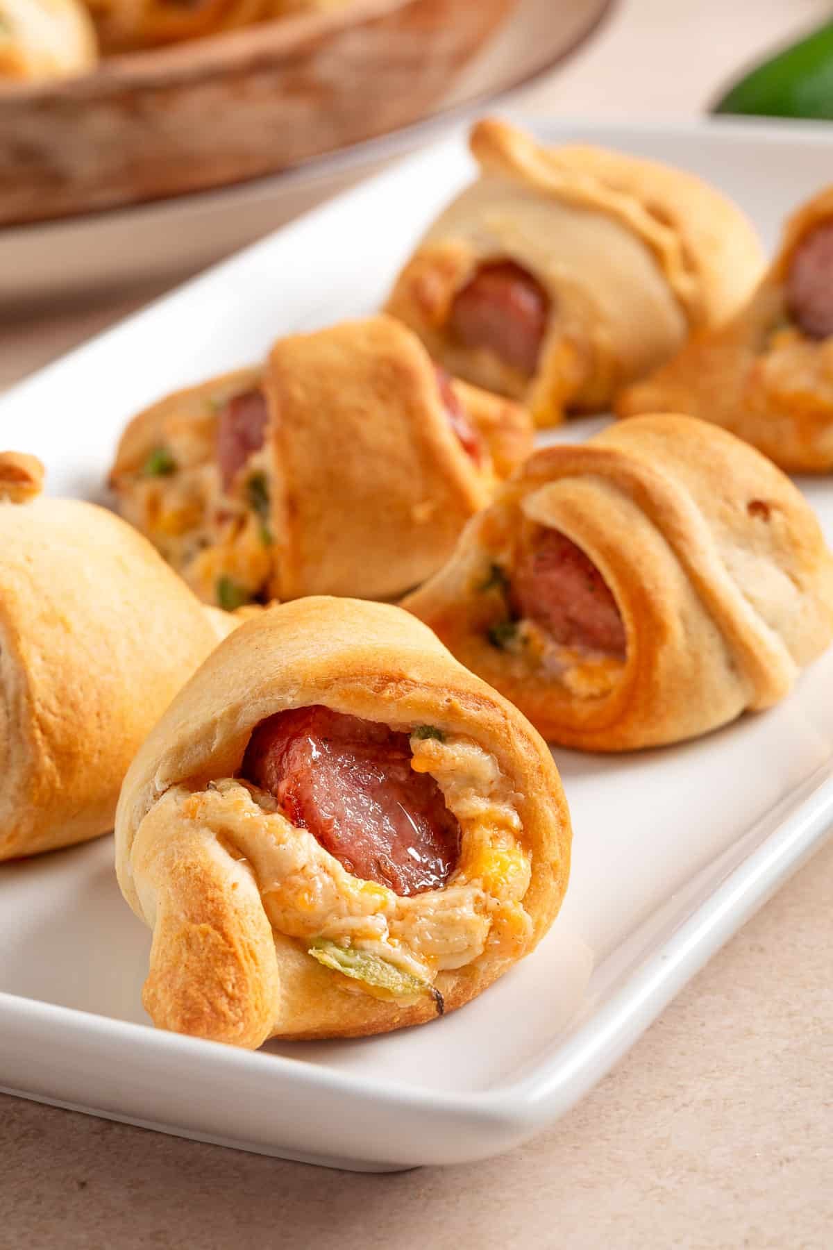 Golden crescent roll-wrapped pigs in a blanket filled with melted cheddar, chopped jalape&ntilde;o, and smoky cocktail sausages, arranged on a white rectangular serving platter.