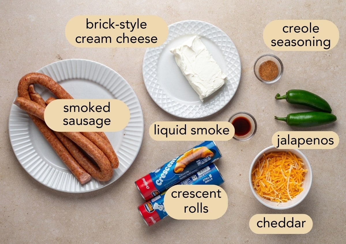 Overhead view of labeled ingredients for jalape&ntilde;o popper pigs in a blanket: smoked sausage links, brick-style cream cheese, shredded cheddar, jalape&ntilde;os, crescent roll tubes, liquid smoke, and Creole seasoning arranged on a beige countertop.