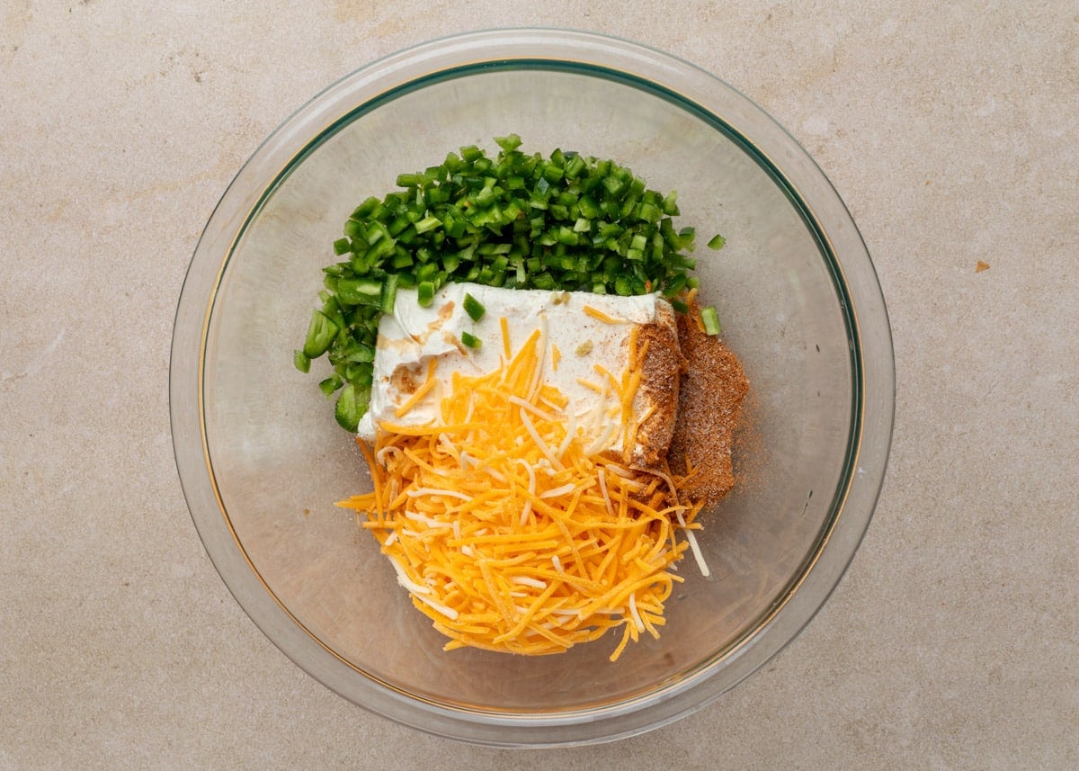 A glass mixing bowl with unblended filling ingredients for Jalape&ntilde;o Popper Pigs in a Blanket, including diced jalape&ntilde;os, brick-style cream cheese, shredded cheddar, and Creole seasoning arranged in separate piles.