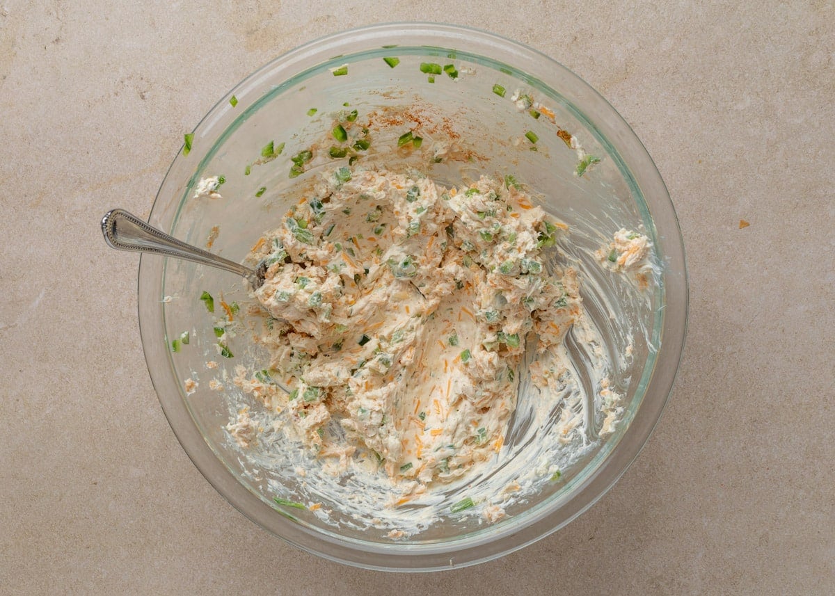 The prepared filling for Jalape&ntilde;o Popper Pigs in a Blanket in a glass bowl, fully mixed into a creamy, spreadable consistency with visible bits of green jalape&ntilde;o and orange cheddar.