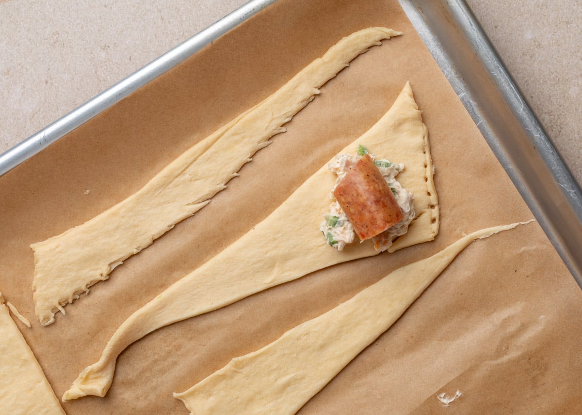 Close-up of crescent roll dough triangle topped with a dollop of jalape&ntilde;o cream cheese mixture and a piece of smoked sausage, ready to be rolled.