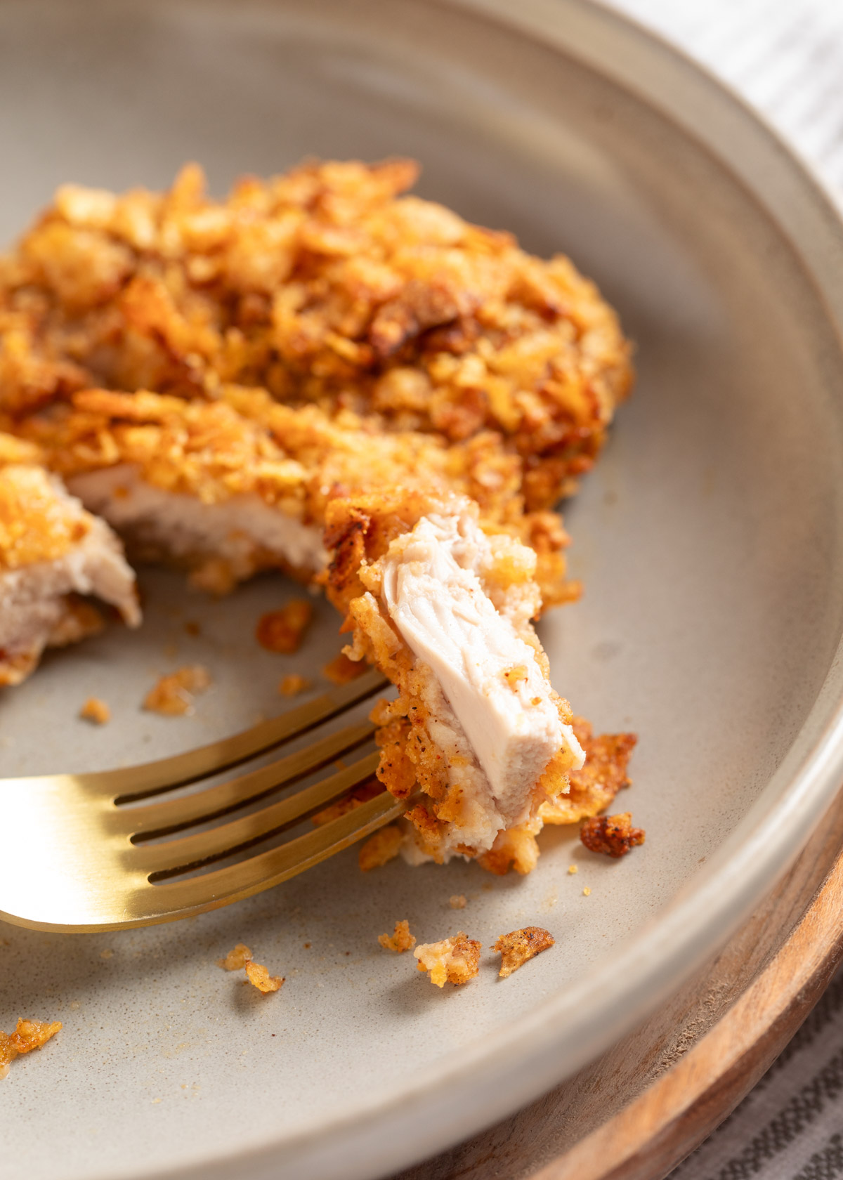 A bite of potato chip-crusted chicken held up by a gold fork, showing the tender, cooked center and craggy, golden-brown coating, with the remaining cutlet resting behind on the plate.