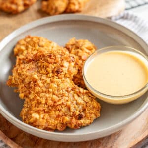Plate of crispy potato chip-coated chicken thighs served with a small bowl of creamy dipping sauce, photographed on a wood charger with a striped napkin in the background.