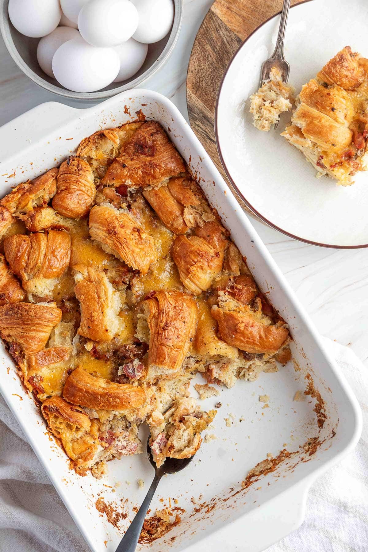 Golden-brown croissant breakfast casserole in a white baking dish with a spoonful missing, showing a flaky, cheesy interior with bacon and sausage. A plated serving sits above the dish next to a bowl of whole eggs.