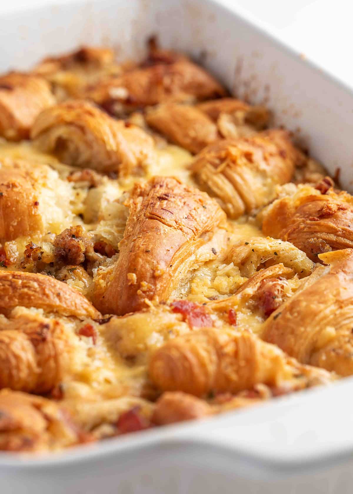 Close-up of a golden-brown baked sausage croissant breakfast casserole in a white dish, showing flaky croissant layers, melted cheese, and crisp bits of bacon and sausage nestled in the custard.