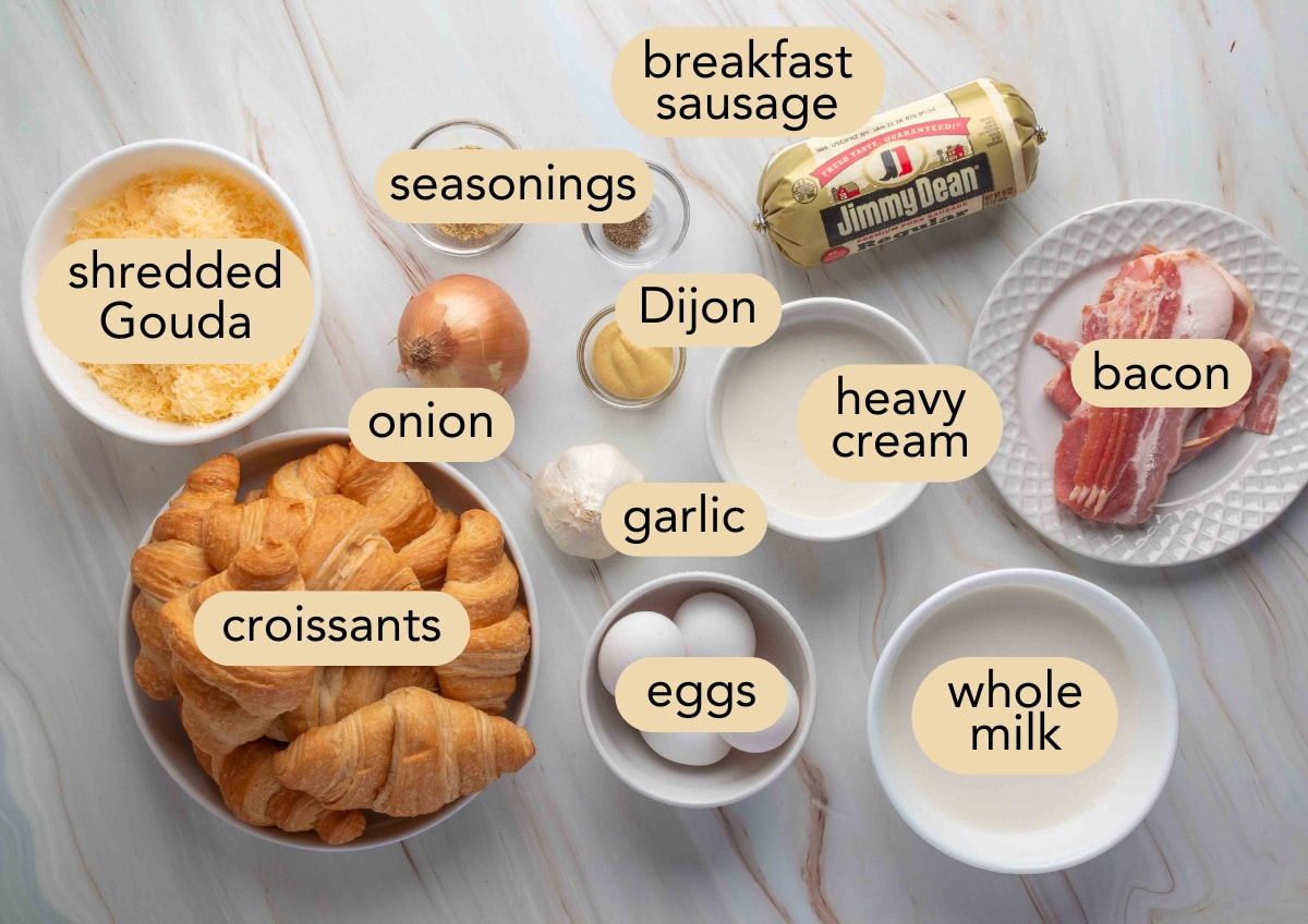 Overhead view of labeled ingredients for a savory breakfast casserole, including croissants, shredded Gouda, breakfast sausage, bacon, onion, garlic, Dijon mustard, eggs, heavy cream, whole milk, and seasonings, all arranged on a light marble surface.