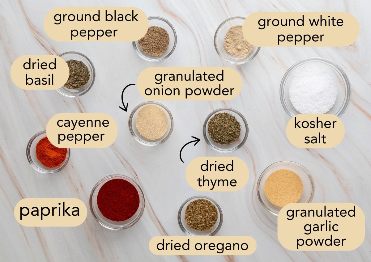 Overhead view of labeled glass bowls showing the ingredients for homemade Creole seasoning, including paprika, cayenne, dried herbs, garlic and onion powders, and kosher salt on a white marble background.