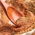 Close-up of a wooden spoon scooping homemade Creole seasoning from a dish, showing a coarse blend of paprika, dried herbs, and salt crystals.