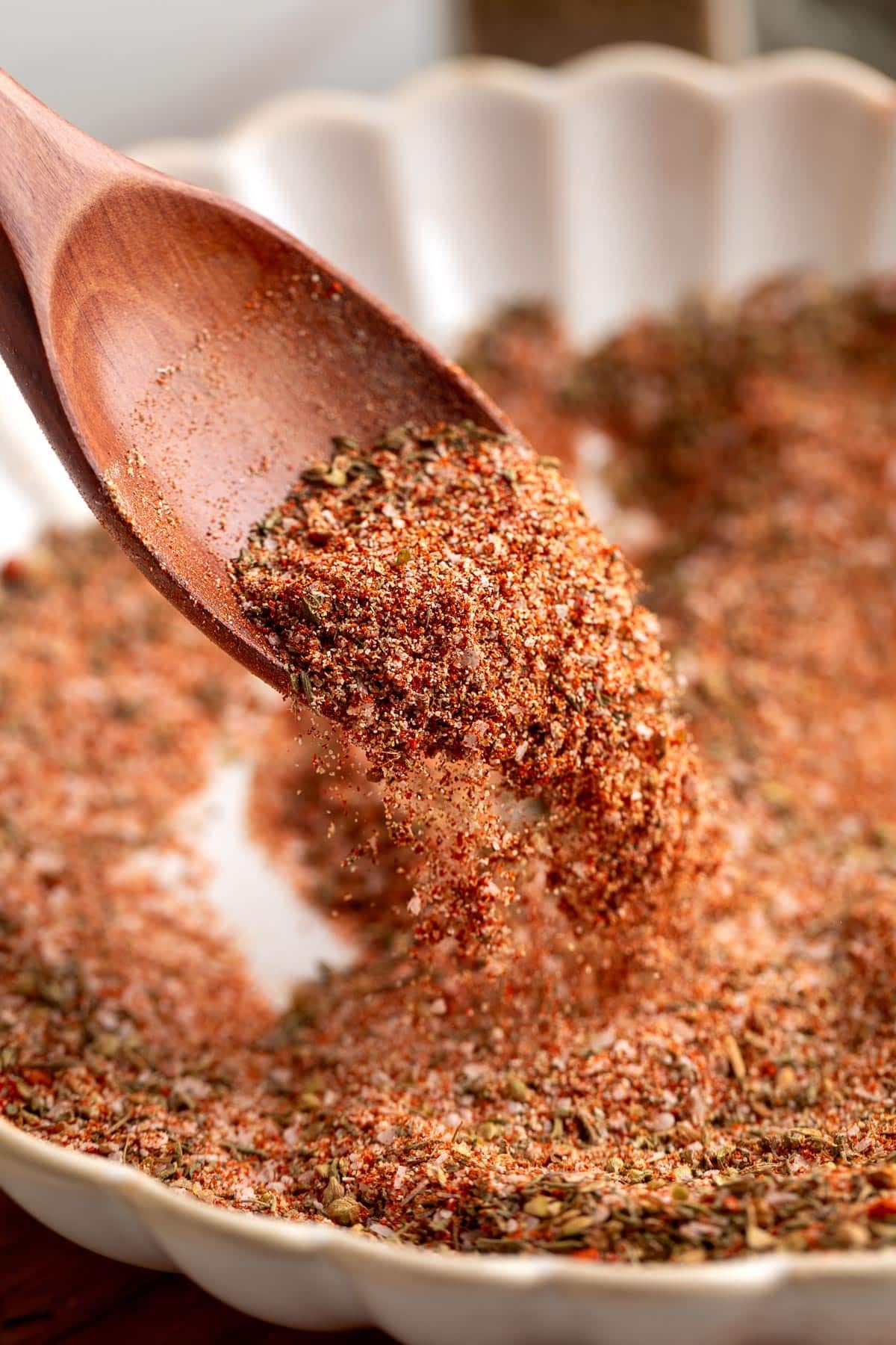 Close-up of a wooden spoon sprinkling a vibrant blend of homemade Creole seasoning into a scalloped white dish, highlighting the mix of herbs and spices.