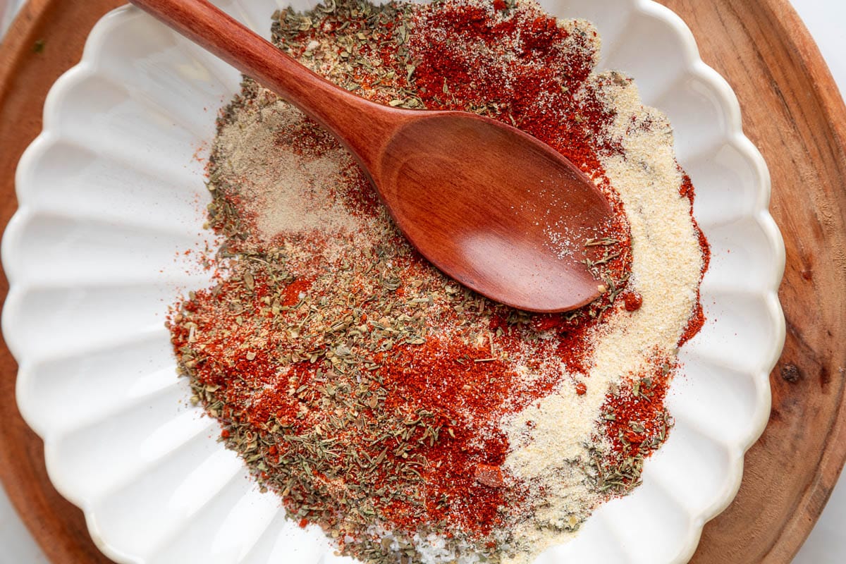 A wooden spoon rests on top of a colorful pile of unmixed dried herbs and spices&mdash;paprika, oregano, garlic powder, and salt&mdash;in a fluted white bowl set on a wooden tray.