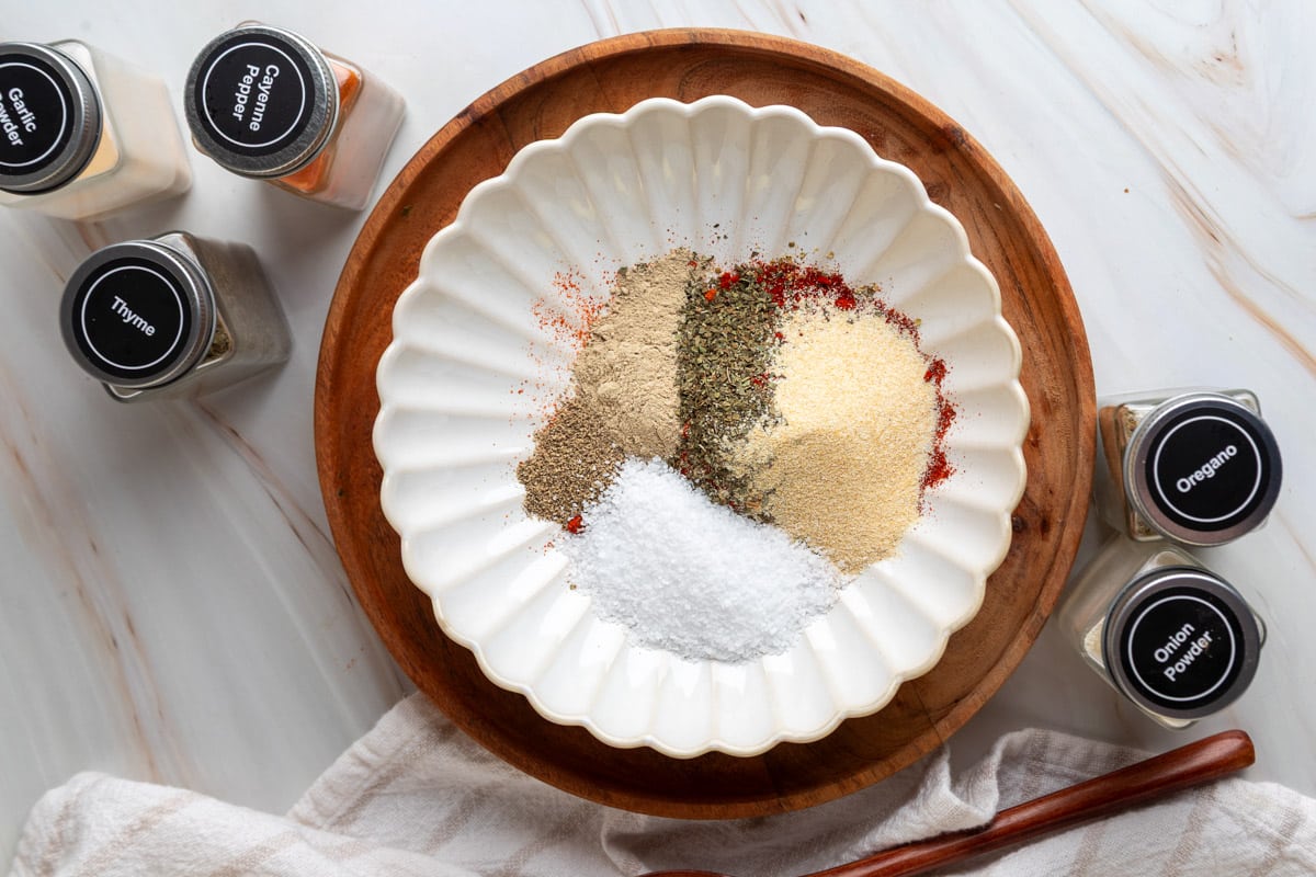 Overhead view of individual spices&mdash;salt, peppers, garlic, onion powder, and herbs&mdash;neatly arranged in a white fluted bowl on a wooden tray, surrounded by labeled spice jars on a marble surface.