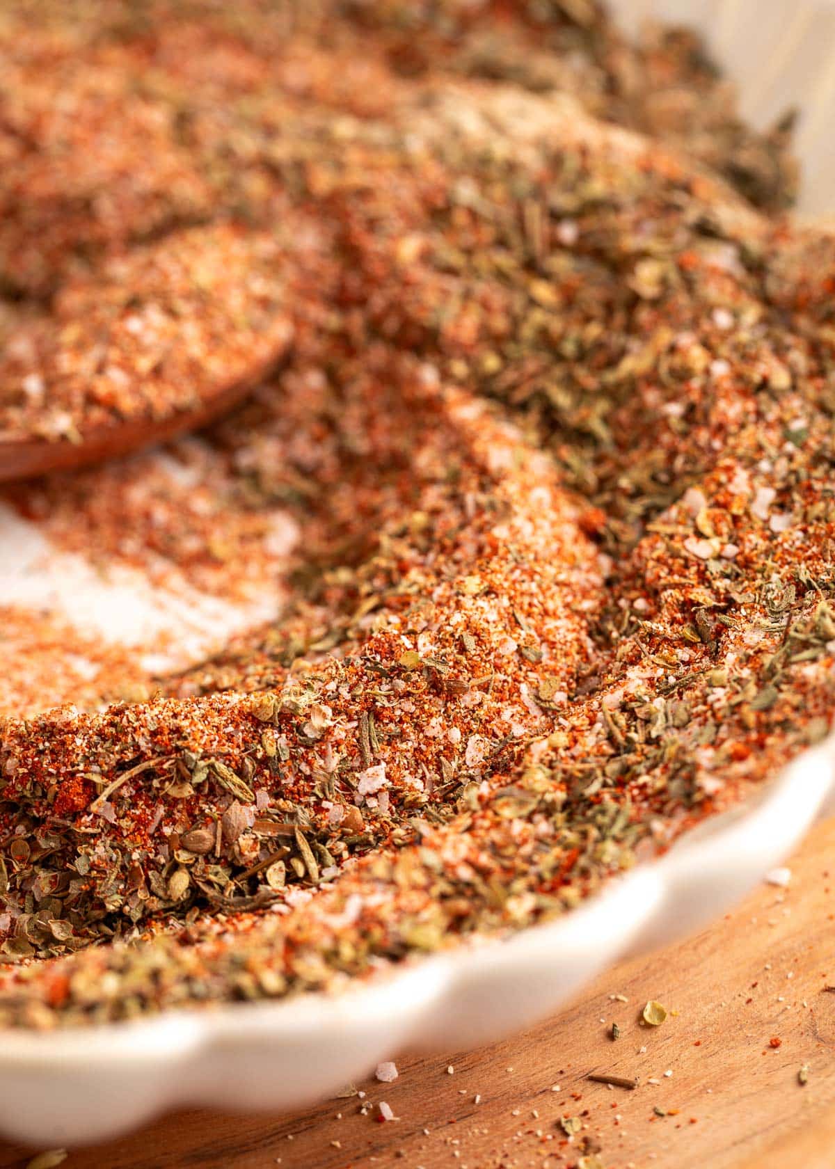 Close-up of a shallow white scalloped dish filled with a homemade Creole seasoning blend, highlighting coarse flakes of dried herbs and red spices with visible salt crystals, photographed on a wooden board with a warm, directional light.