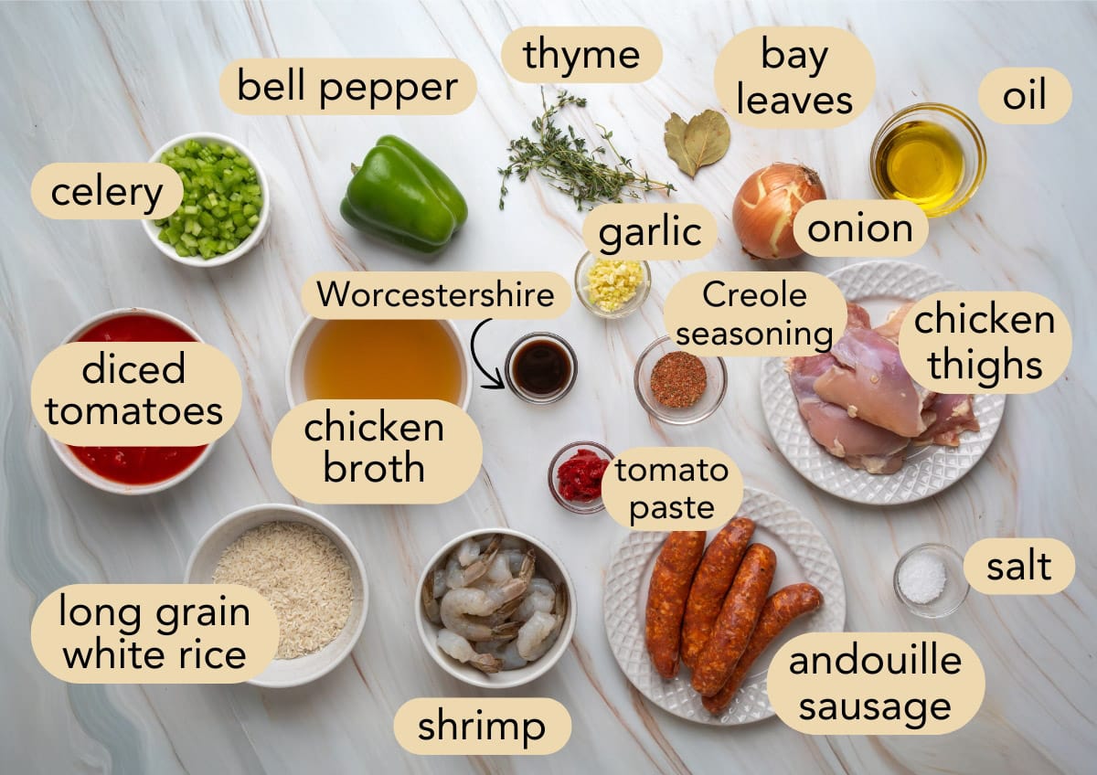 Overhead view of labeled ingredients for jambalaya soup arranged on a marble surface, including shrimp, andouille sausage, chicken thighs, diced tomatoes, rice, vegetables, herbs, spices, broth, and seasonings.