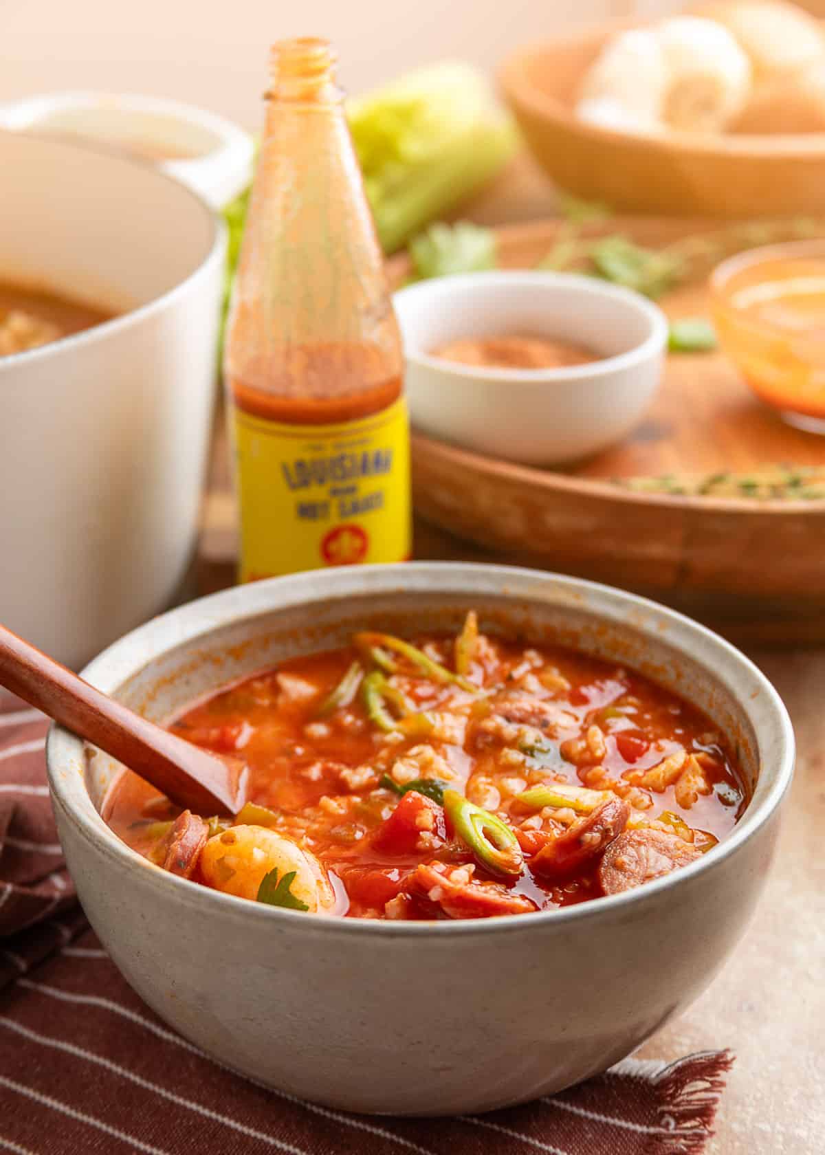 A ceramic bowl filled with jambalaya soup sits on a striped cloth napkin, topped with sliced green onions. A bottle of Louisiana hot sauce and fresh ingredients are softly blurred in the background.