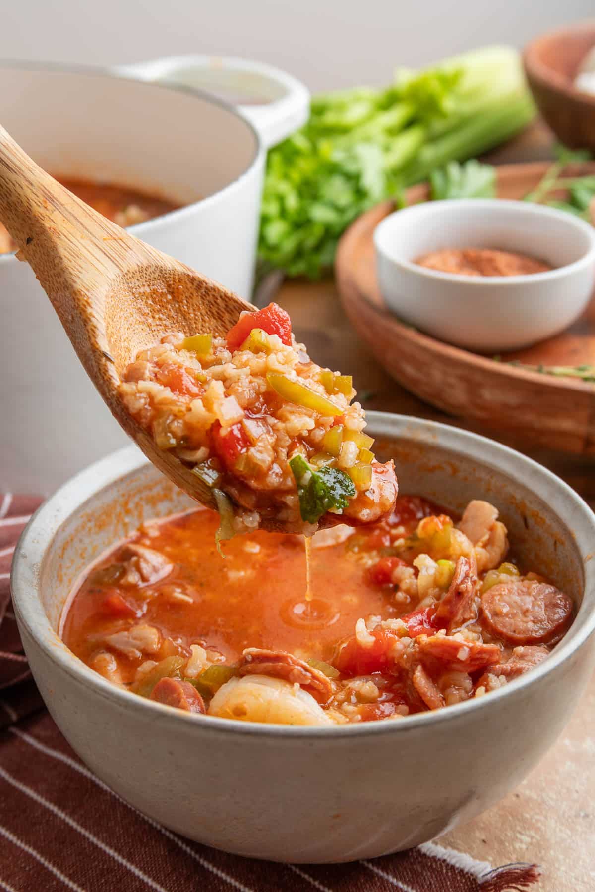 A wooden spoon holds a hearty scoop of jambalaya soup above a bowl, showcasing colorful vegetables, rice, sausage, and herbs. A Dutch oven of soup and fresh herbs are blurred in the background.