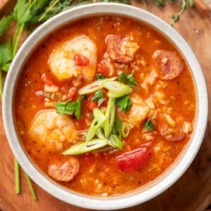 Overhead view of a bowl of jambalaya soup filled with shrimp, sausage, tomatoes, and rice, topped with sliced green onions and chopped parsley. Fresh herbs rest on a wooden surface in the background.