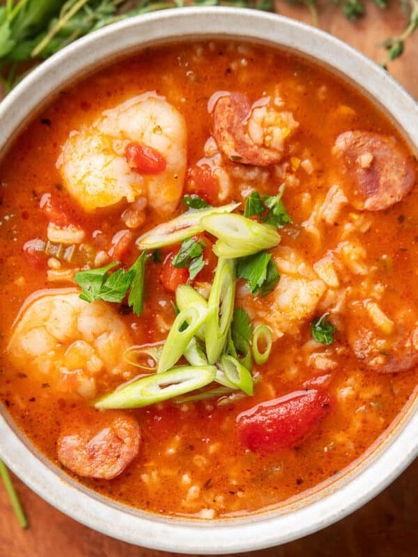 Overhead view of a bowl of jambalaya soup filled with shrimp, sausage, tomatoes, and rice, topped with sliced green onions and chopped parsley. Fresh herbs rest on a wooden surface in the background.