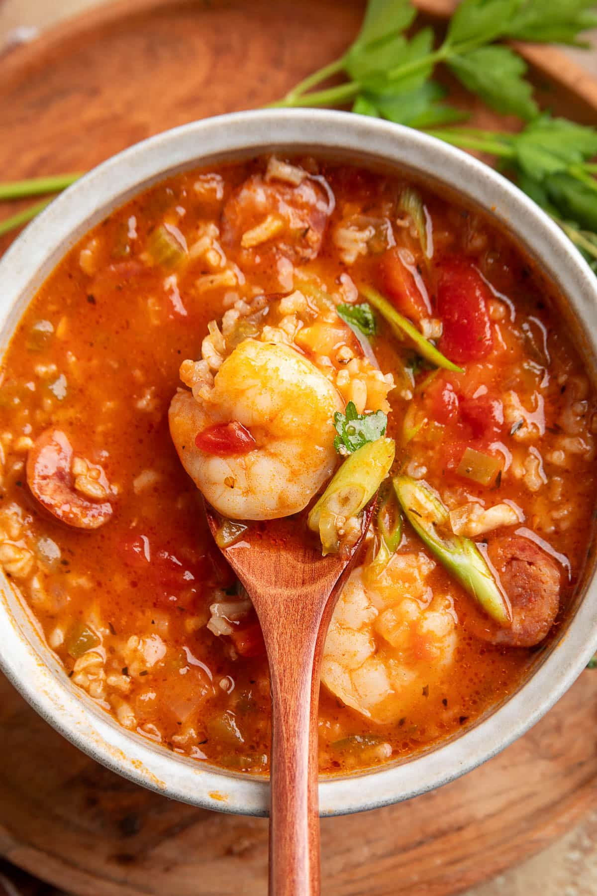 Close-up of a wooden spoon lifting a shrimp from a bowl of jambalaya soup, surrounded by sausage, rice, tomatoes, and sliced green onions in a spicy, red broth. Fresh parsley rests in the background.