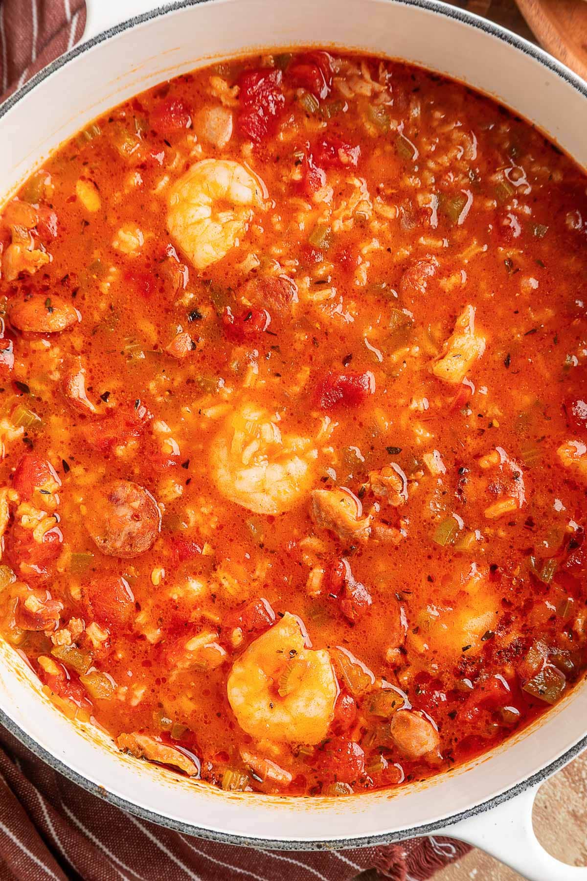 Overhead view of a Dutch oven filled with jambalaya soup, showcasing a vibrant tomato-based broth with shrimp, sliced sausage, chicken, rice, and peppers.