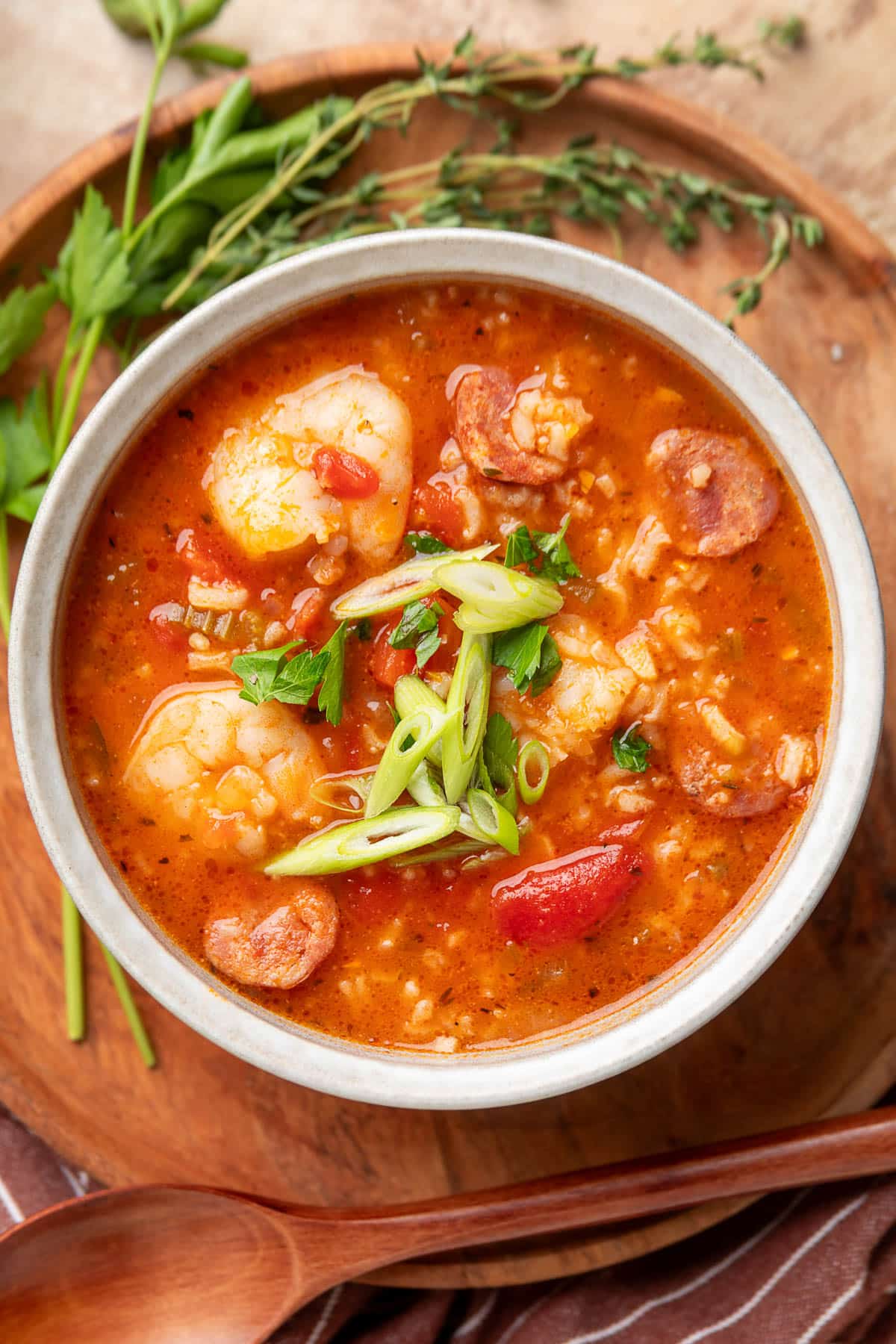 Overhead view of a bowl of jambalaya soup with shrimp, andouille sausage, tomatoes, and rice, topped with sliced scallions and parsley, served on a wooden tray with fresh herbs in the background.