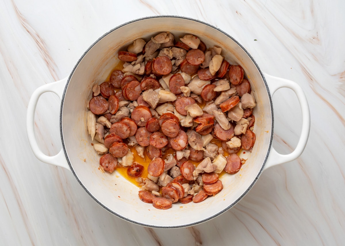 Overhead view of a white Dutch oven filled with browned slices of andouille sausage and chunks of chicken thighs, showing rendered drippings at the bottom of the pan.