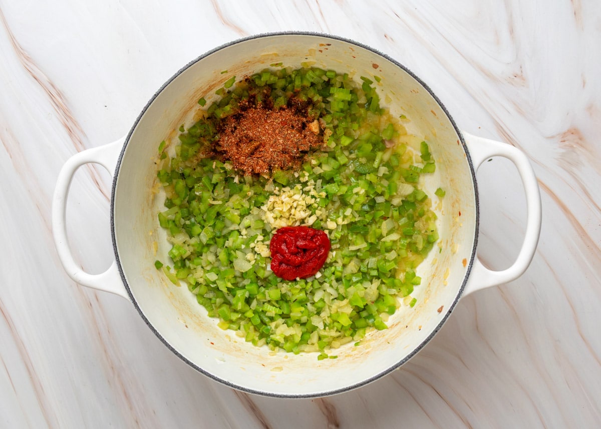 Uncooked chopped celery, onion, and bell pepper in a white Dutch oven with tomato paste, minced garlic, and Creole seasoning added on top before being stirred together.