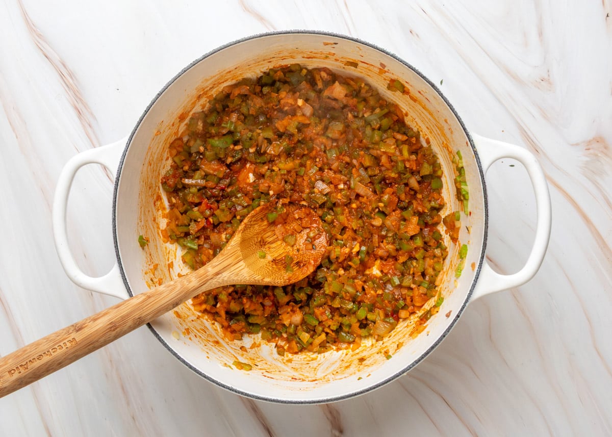 Cooked-down mixture of celery, onion, bell pepper, tomato paste, garlic, and Creole seasoning in a white Dutch oven with a wooden spoon resting in the pot.