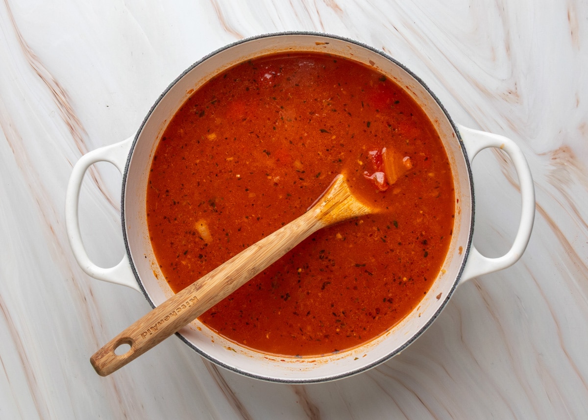 Red, well-seasoned broth with visible herbs, tomato chunks, and a few pieces of meat simmering in a white Dutch oven, stirred with a wooden spoon.