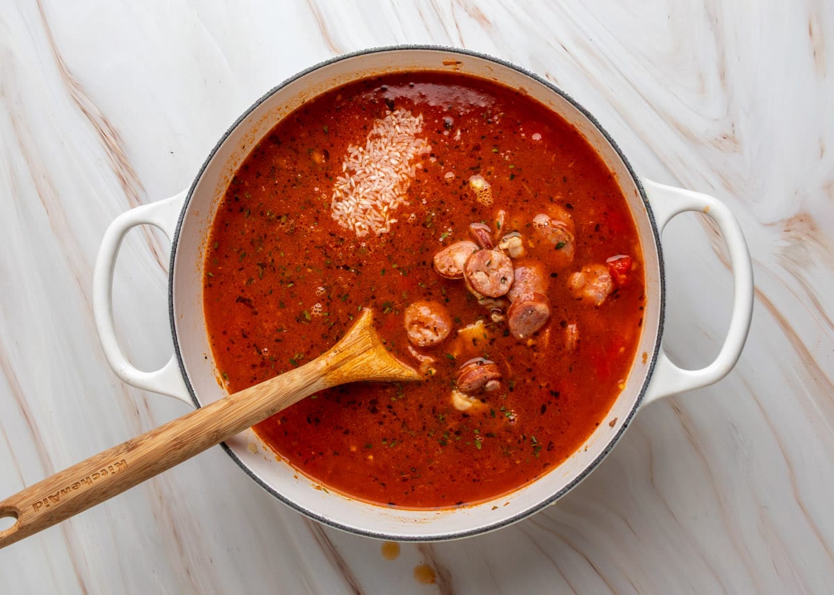 Uncooked white rice and browned sausage slices are added to a rich, red tomato-based broth in a white Dutch oven, with a wooden spoon resting in the pot.