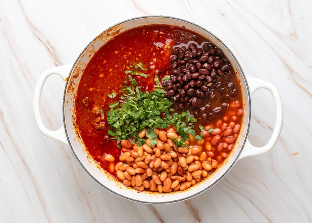 Overhead photo of a pot of chili with visible layers of black beans, pinto beans, kidney beans, and chopped fresh cilantro just added to a vibrant red broth, ready to be stirred together.