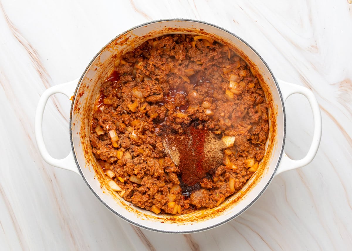 Overhead view of a Dutch oven filled with saut&eacute;ed ground meat and onions, with a mound of chili spices including paprika, cumin, and chipotle powder added to the center before stirring.