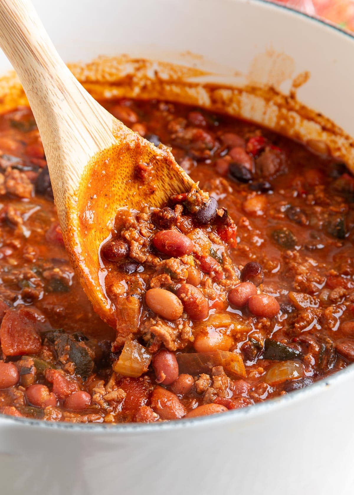 A wooden spoon lifts a scoop of spicy chili from a white Dutch oven, highlighting the beans, ground meat, and chunks of tomato in a vibrant, spiced broth.