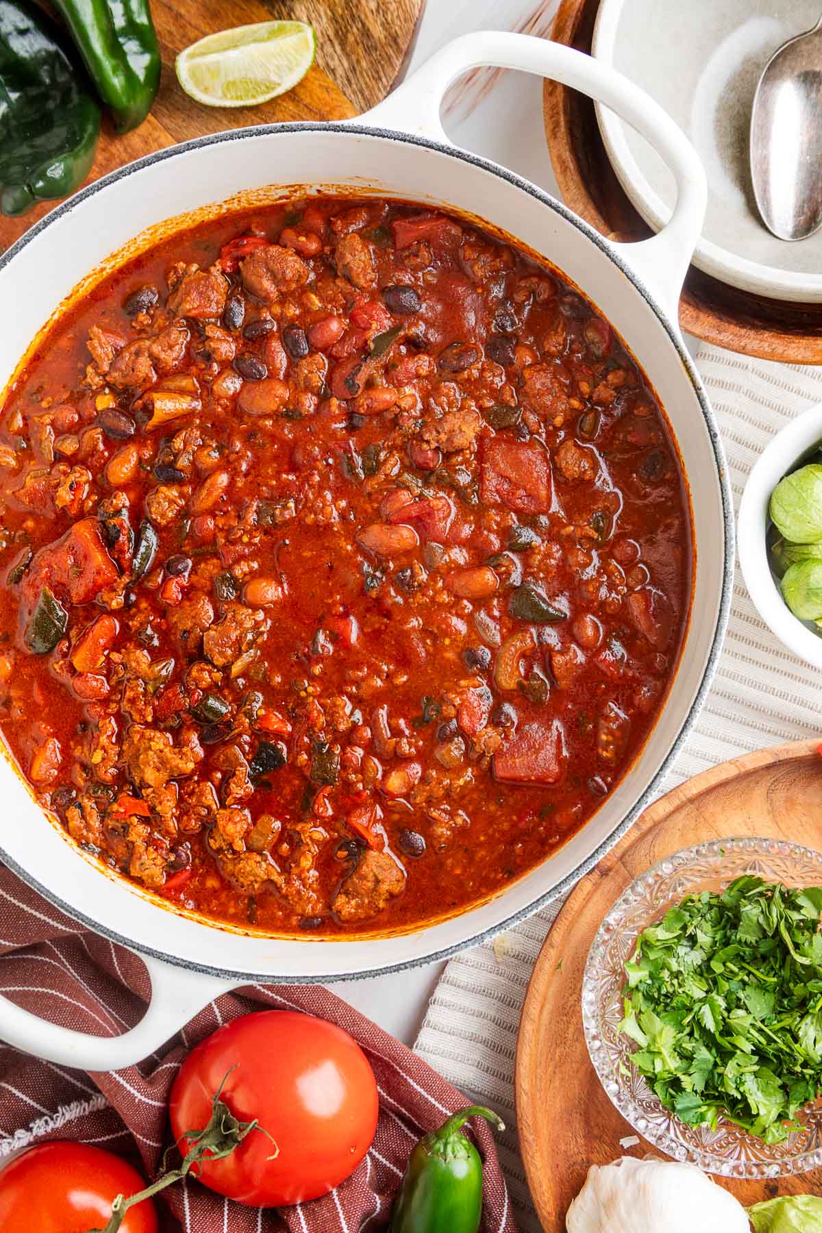 Overhead view of a white Dutch oven filled with hearty chili, surrounded by fresh ingredients including tomatoes, jalape&ntilde;o, cilantro, garlic, and a halved lime, styled on a striped cloth and wood cutting boards.