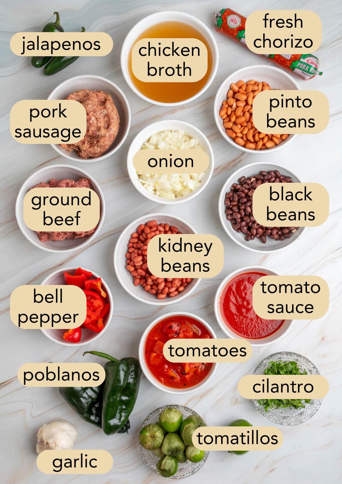 Overhead view of labeled chili ingredients in bowls and on the counter, including three types of beans, ground meats, chopped onions, bell peppers, tomatoes, garlic, tomatillos, and fresh herbs.