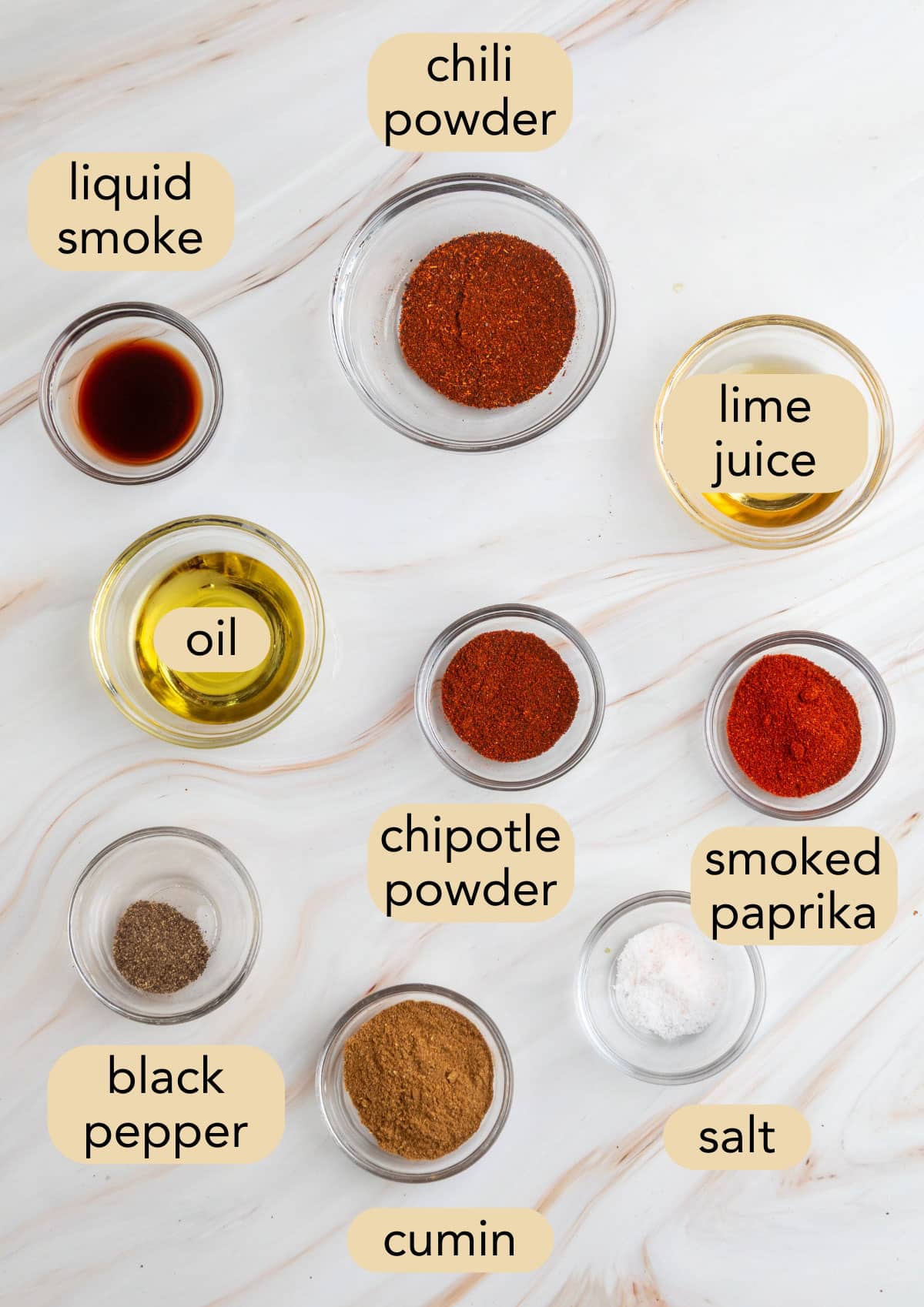 Overhead view of labeled chili seasonings in small glass bowls, including chili powder, chipotle powder, smoked paprika, cumin, black pepper, salt, oil, lime juice, and liquid smoke.